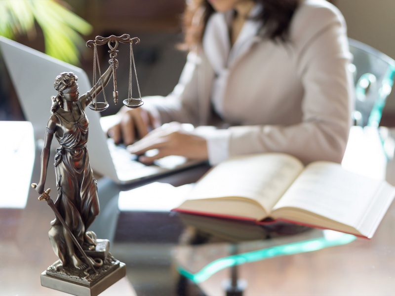 Woman typing on a laptop with a book open beside her and the scales of justice.