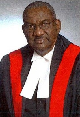 Bald black man with glasses wearing red, white and black judicial robes. 