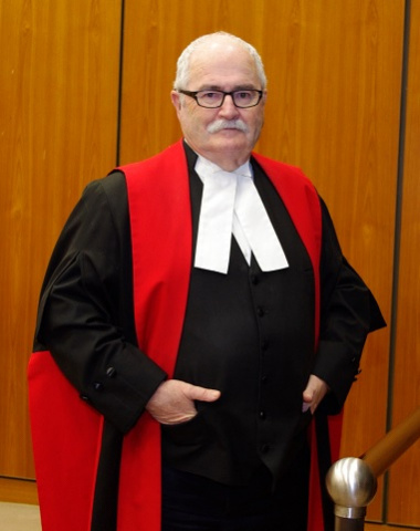 White man with glasses wear black, red and white judicial robes and his hands in his pockets.