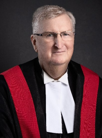 White man wearing glasses and red, black and white judicial robes. 