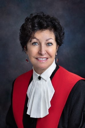 Female judge with short dark hair, wearing black and red judicial robes with a lacy collar. 