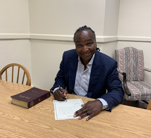 Black man in a suit jacket seated at a table signing a document. 