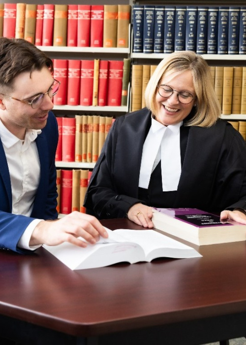 A female judge in black robes and a man in a blue suit sit at a desk in front of an open laptop. Behind them are shelves of legal books. 