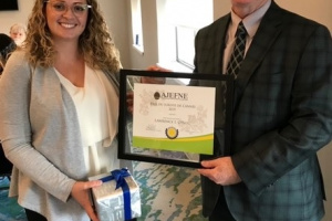 White man wearing glasses and a suit accepts a framed award from a woman. 