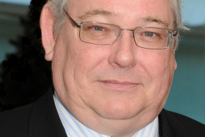 Man with white hair and glasses wearing a suit and checkered tie. 