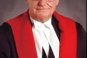 White man with white hair and glasses wearing black, red and white judicial robes. 