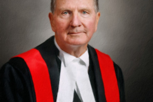 Photo of a male judge wearing black and white judicial robes with a red sash. 